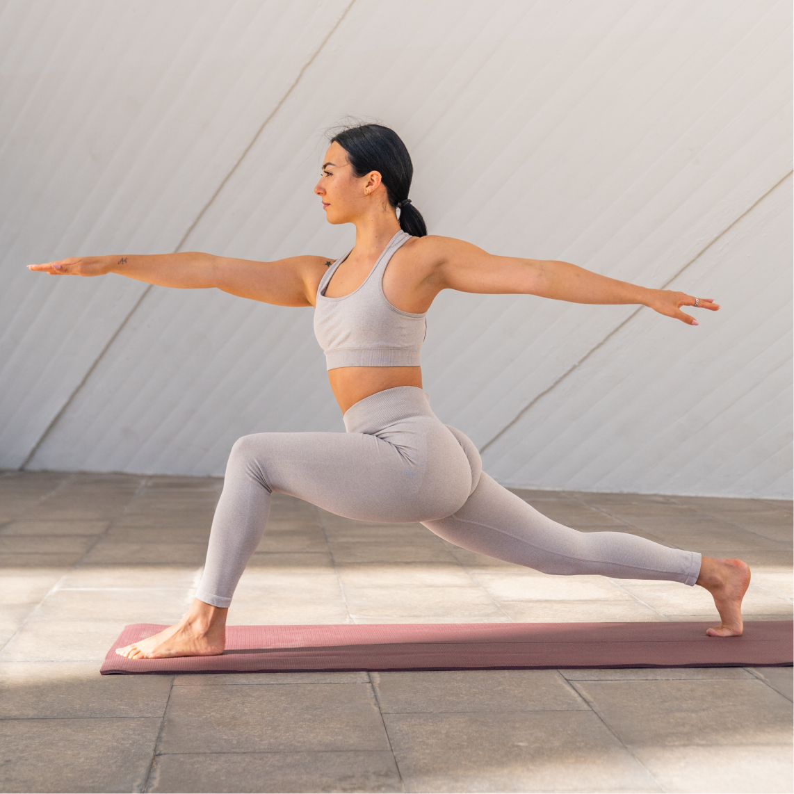 A woman in light grey activewear practises yoga indoors, performing a Warrior II pose on a mauve mat. Perfect for fitness case studies, the minimal and bright background highlights her strong form as she gazes over her right hand.