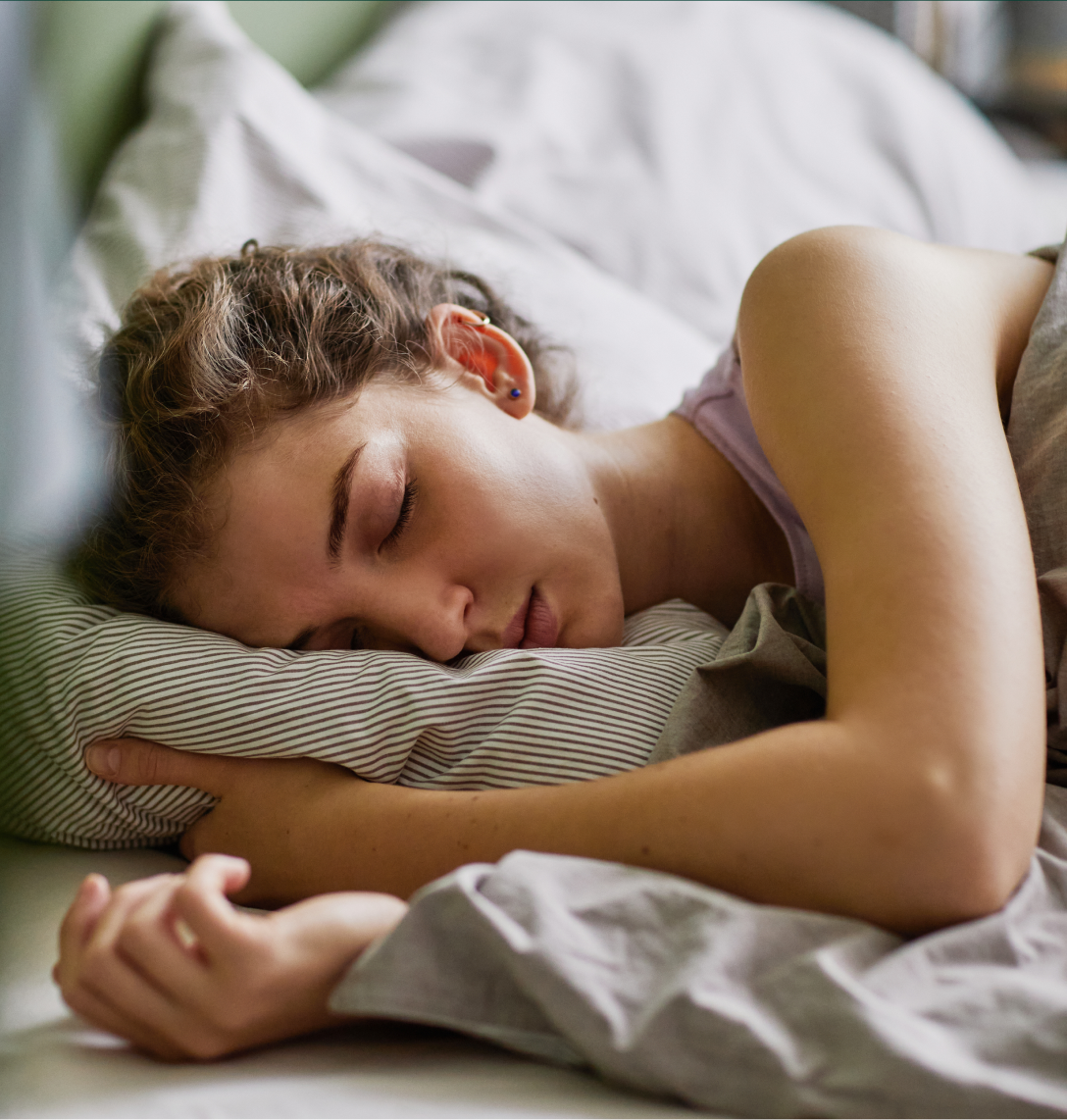 A young woman with curly brown hair sleeps on her side in bed, resting her head on a striped pillow—like a peaceful subject in case studies exploring restful sleep, she’s covered with a greyish blanket and appears completely relaxed.