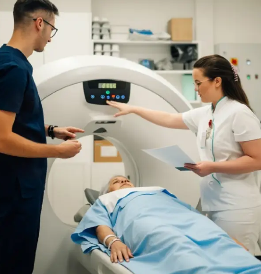 Two healthcare professionals stand beside a patient lying on a hospital bed, preparing for a CT scan. The staff adjust controls and review case studies in a bright, modern medical room as the patient waits in a blue gown.