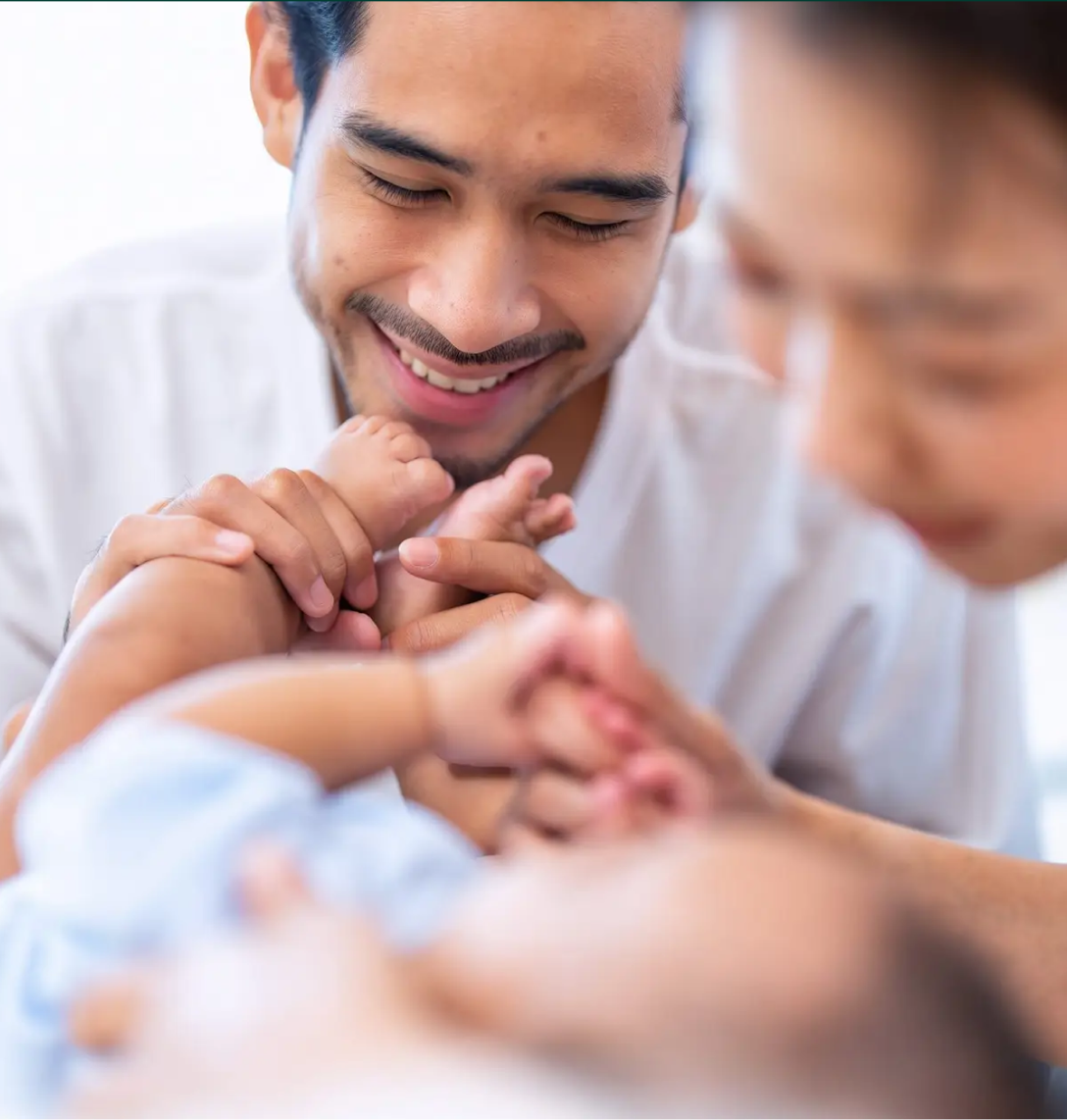 A smiling man gently holds a baby’s feet, looking down lovingly. A woman, blurred in the foreground, also interacts with the baby. The warm, soft-lit scene beautifully captures family bonding—much like case studies highlight meaningful connections.