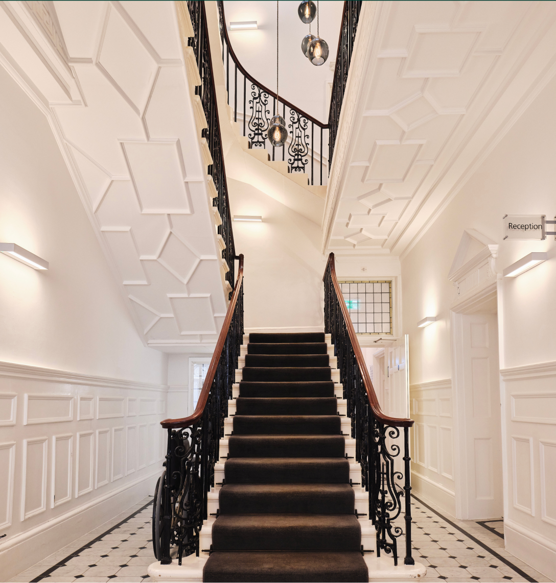 Grand staircase with a black runner carpet, ornate black railings, and white panelled walls. Geometric ceiling patterns, modern lights, and a tiled black-and-white floor set the scene—perfect for showcasing inspiring case studies. Reception sign visible on the right wall.
