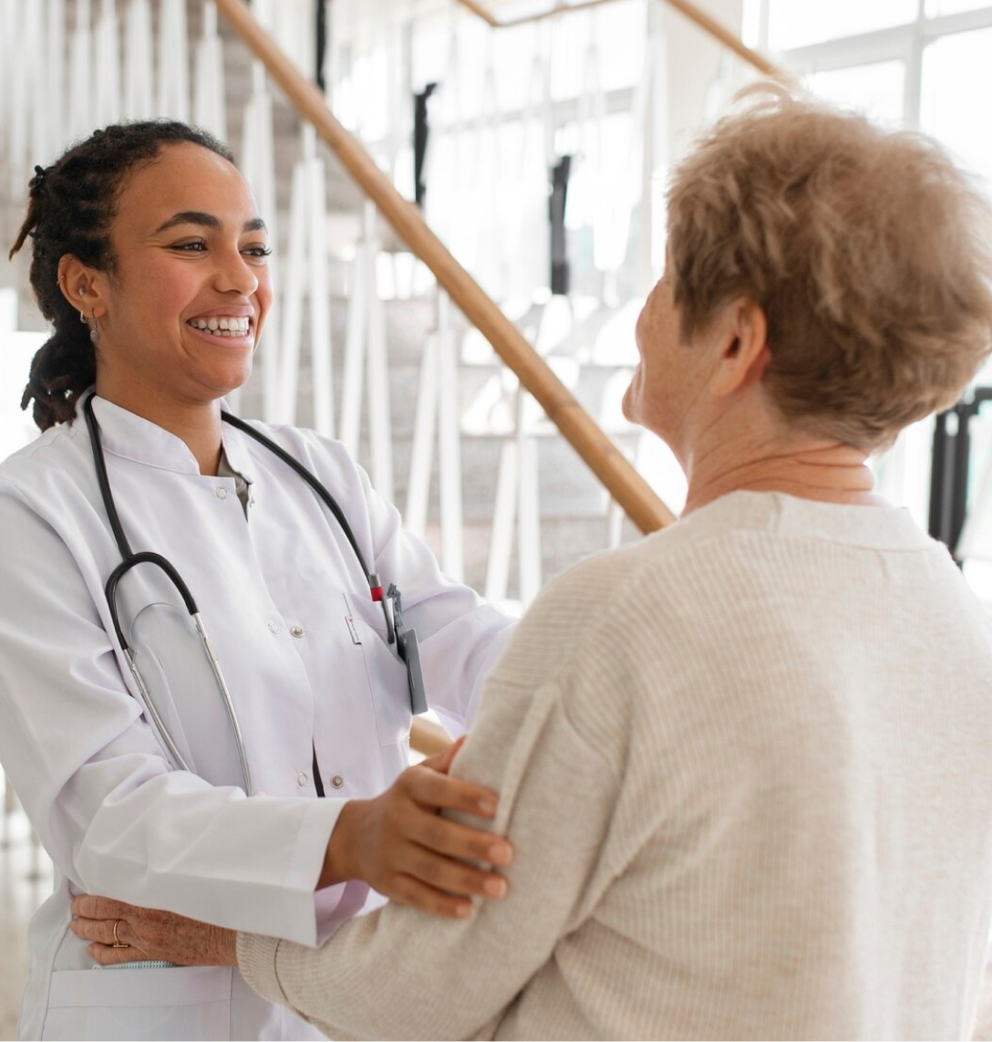 A smiling female doctor in a white coat and stethoscope warmly greets an older woman, exemplifying compassionate care often highlighted in case studies, both standing indoors near a staircase with bright natural light.