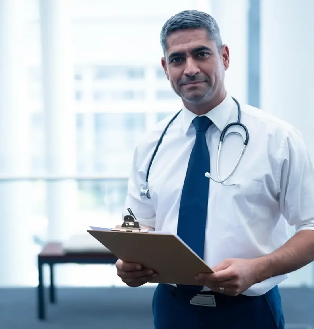 A male doctor with short grey hair, wearing a white shirt, blue tie, and stethoscope around his neck, holds a clipboard of case studies and smiles softly in a bright, modern medical office with large windows in the background.