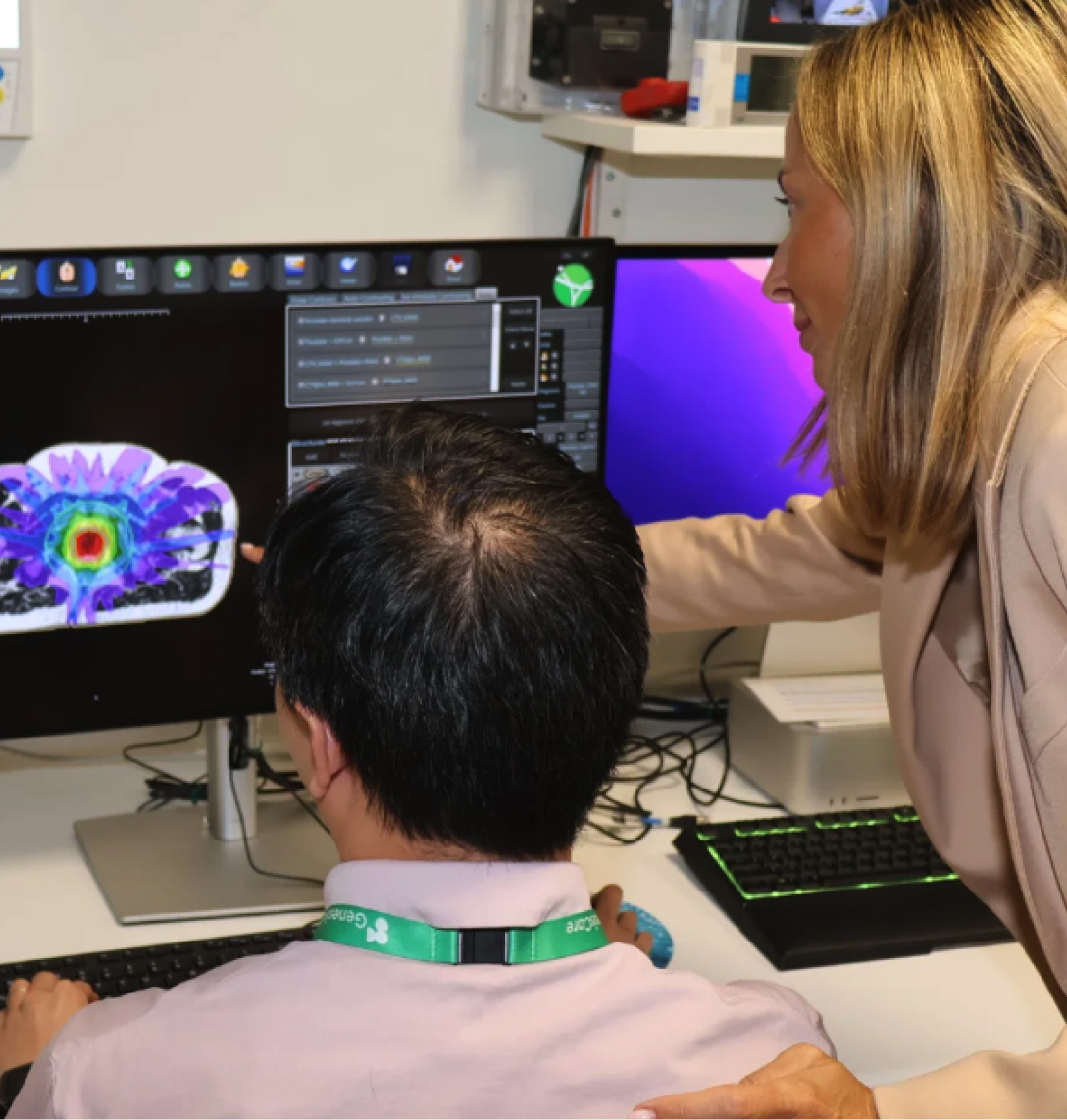 A woman in a beige blazer stands beside a seated man, pointing at a monitor displaying colourful case studies from medical scans. The man, wearing a white shirt and green lanyard, looks at the screen in an office with computers and medical equipment.