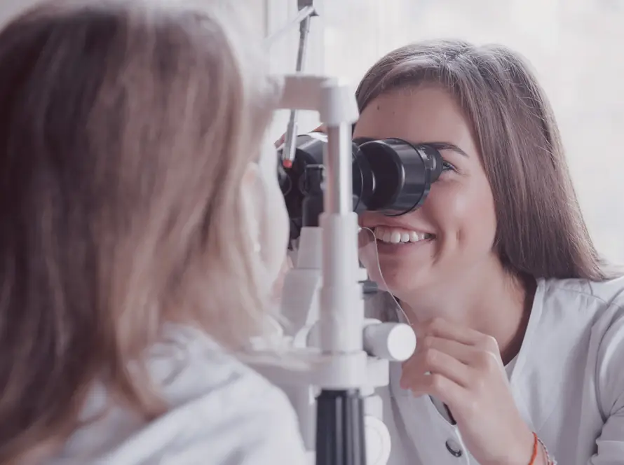 A smiling female optometrist examines a patient’s eyes using a slit lamp in a bright room. The patient, with long light brown hair, sits facing the device whilst the optometrist looks through the microscope, focusing on the patient’s eye.
