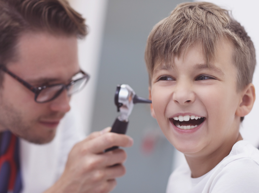 A smiling boy sits while a doctor examines his ear with an otoscope. The doctor wears glasses and a white coat. The boy looks happy and relaxed in a brightly lit medical surgery. The background is blurred, focusing on their interaction.