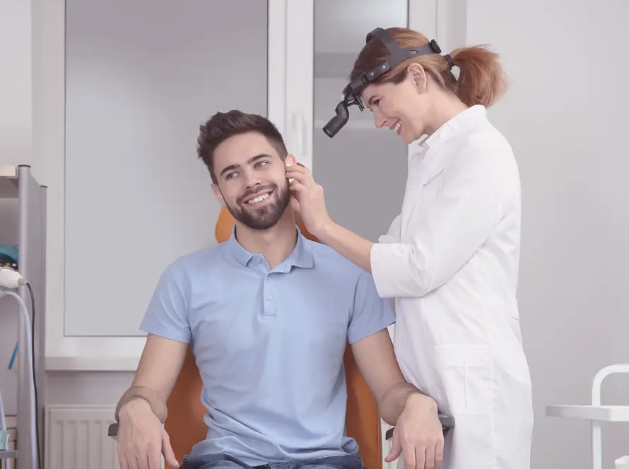 A smiling male patient sits in a clinic chair whilst a female doctor in a white coat and head torch examines his ear. Medical equipment is visible around them, and the setting is a modern, bright examination room. Both appear relaxed and comfortable.