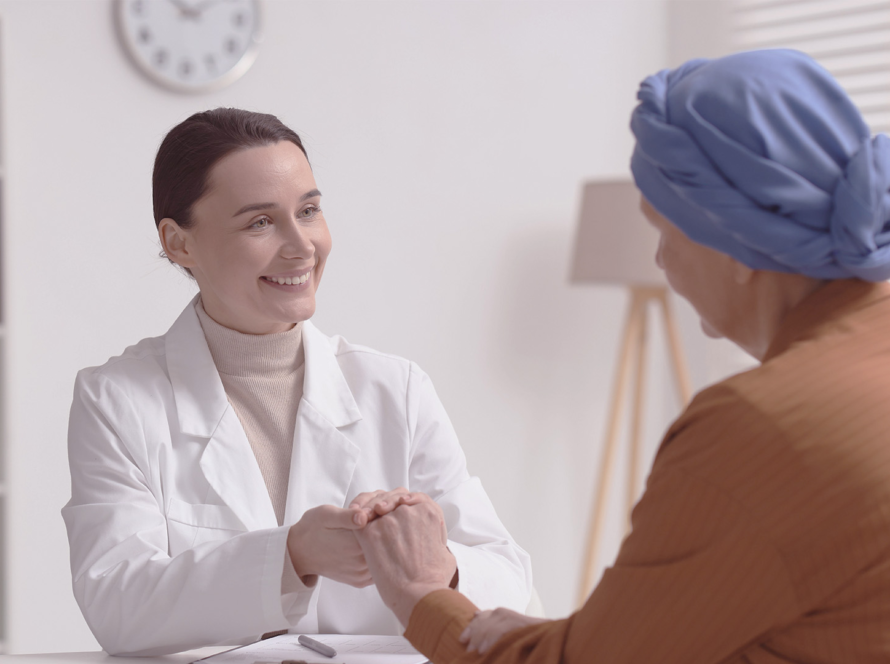 A female doctor in a white coat warmly holds the hands of a patient wearing a blue headscarf and brown top. They are seated across a desk in a bright office, suggesting a supportive medical consultation. A clock and shelves are visible in the background.