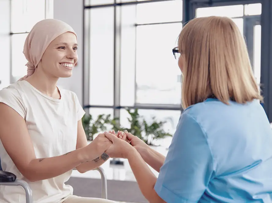 A smiling woman wearing a headscarf sits in a wheelchair, holding hands with a healthcare professional in blue scrubs. They face each other in a bright room with large windows and plants, suggesting support and encouragement.
