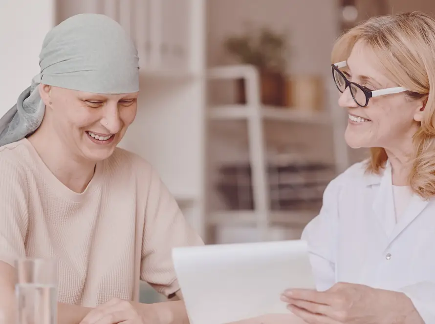 A woman with a light headscarf, appearing as a cancer patient, smiles whilst sitting with a female doctor in a white coat who is holding and discussing a document. Both women look happy and engaged in a bright, comforting room.