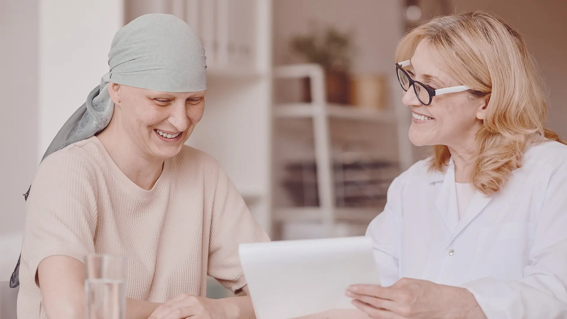 A woman with a light headscarf, appearing as a cancer patient, smiles whilst sitting with a female doctor in a white coat who is holding and discussing a document. Both women look happy and engaged in a bright, comforting room.