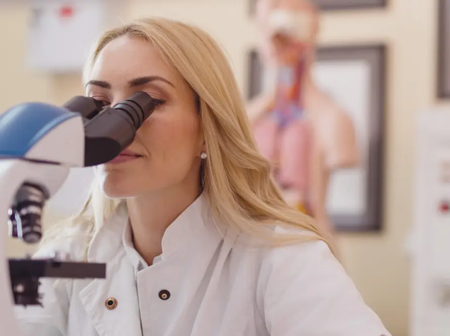 A woman with long blonde hair wearing a white lab coat looks into a microscope in a laboratory setting. Anatomical models and scientific posters are blurred in the background, indicating a medical or research environment.