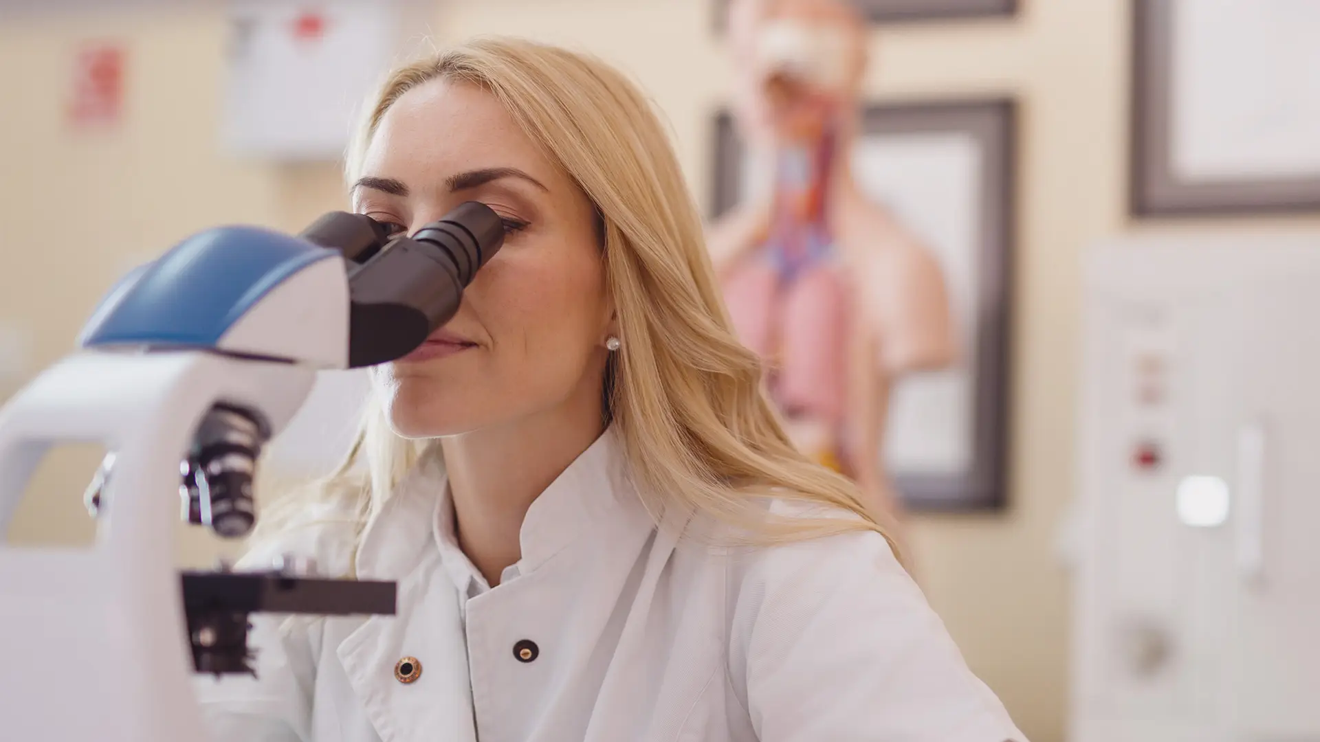 A woman with long blonde hair wearing a white lab coat looks into a microscope in a laboratory setting. Anatomical models and scientific posters are blurred in the background, indicating a medical or research environment.