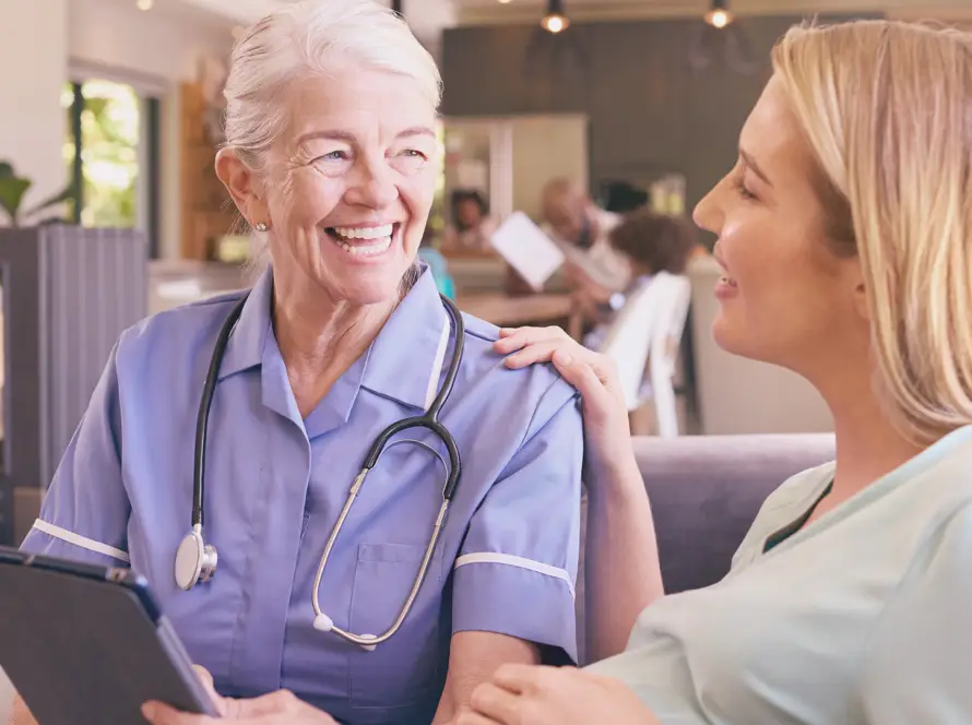 An older female nurse with short grey hair, wearing a blue uniform and stethoscope, smiles warmly whilst holding a clipboard and talking to a younger woman with blonde hair, who gently touches the nurse’s shoulder. They sit in a bright, cosy room.