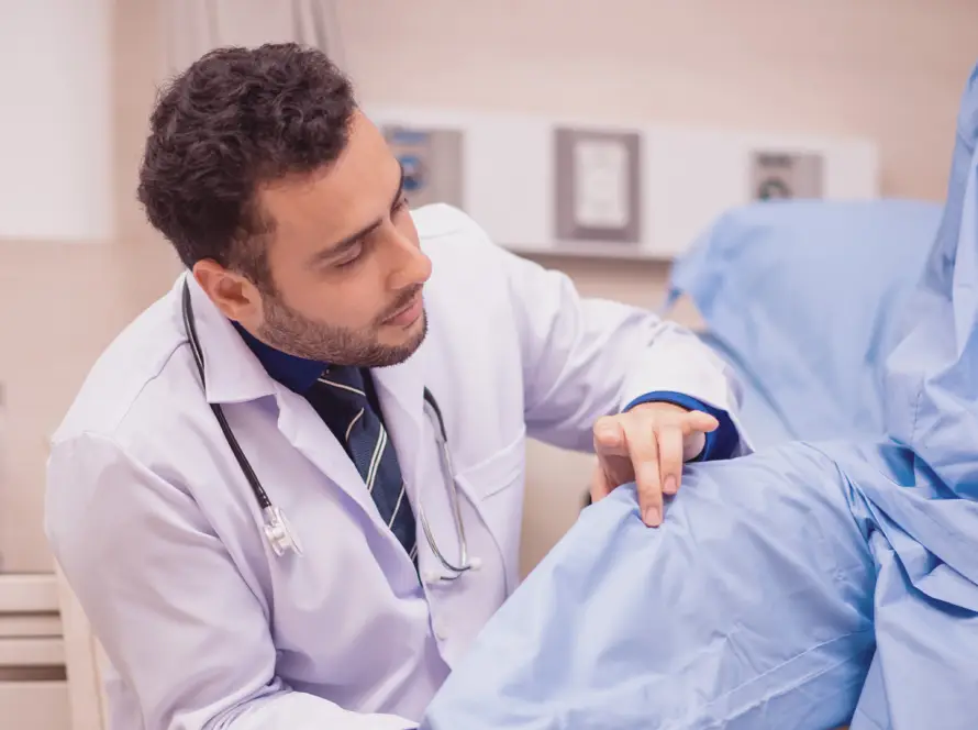 A male doctor in a white coat examines a patient’s knee in a medical setting. The doctor is focused, gently touching the patient’s covered leg while the patient sits on an examination bed wearing a blue gown. Medical equipment is visible in the background.
