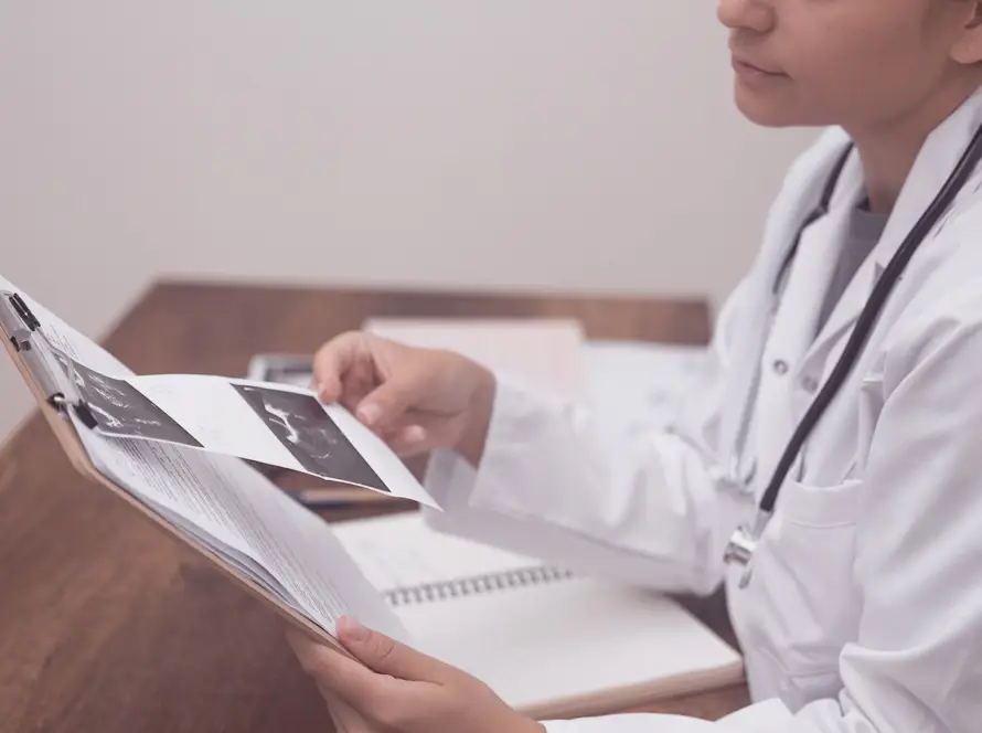A doctor in a white coat sits at a desk holding a clipboard and examining ultrasound images. The desk has an open notebook and some papers. The doctor is partially visible from the side, with a stethoscope round their neck.