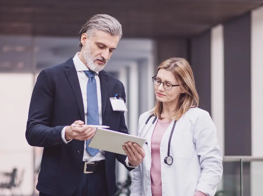 A man in a suit holds a tablet and discusses its contents with a woman in a white coat and stethoscope. They stand indoors, appearing focused and engaged in conversation, possibly collaborating in a professional or healthcare setting.