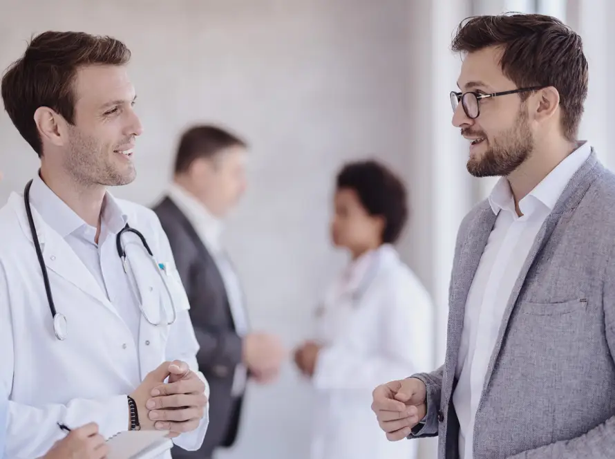 Three healthcare professionals, two in white coats with stethoscopes and one in scrubs, talk with a man in a suit jacket. Two more people converse in the blurred background. The group stands in a bright, modern medical practice.