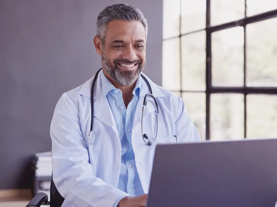 A smiling male doctor with grey hair and beard, wearing a white lab coat and stethoscope, sits at a desk using a laptop. Large windows behind him let in natural light, and he appears focused and approachable in a modern office setting.