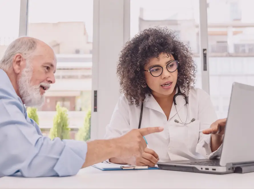 A female doctor with curly hair and glasses sits at a desk, showing information on a laptop to an older male patient with a beard. Both are engaged in discussion, gesturing towards the screen, with large windows and buildings in the background.