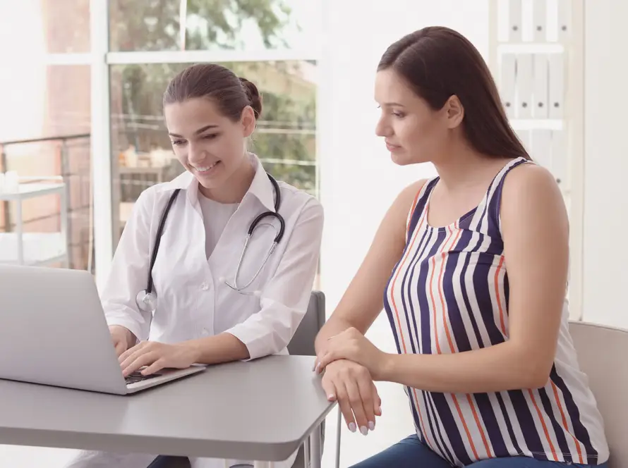 A female doctor in a white coat and stethoscope sits at a desk with a laptop, smiling whilst showing something on the screen to a seated woman in a striped sleeveless top. They are in a bright medical surgery with shelves and large windows.