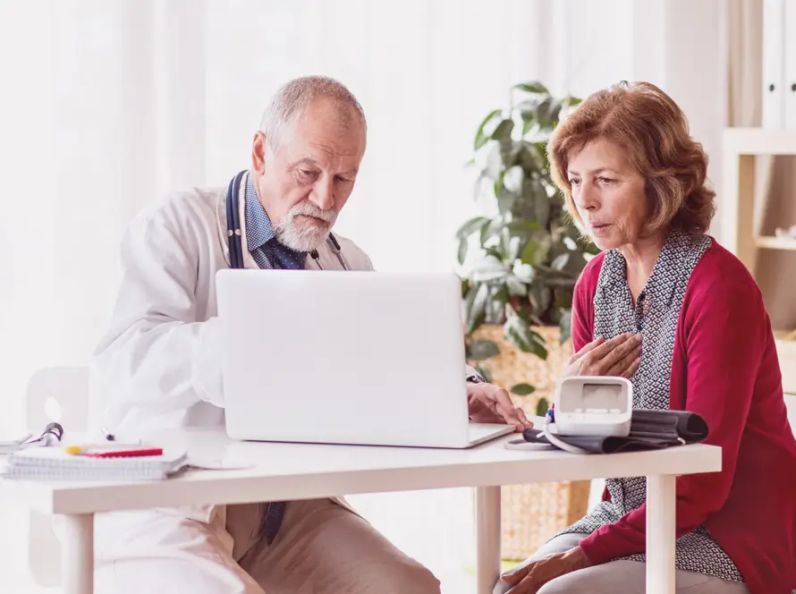 An older male doctor shows information on a laptop to an older woman, who looks concerned and touches her chest. They sit at a white desk with a blood pressure monitor and notepad in a bright, modern medical surgery with plants and shelves.
