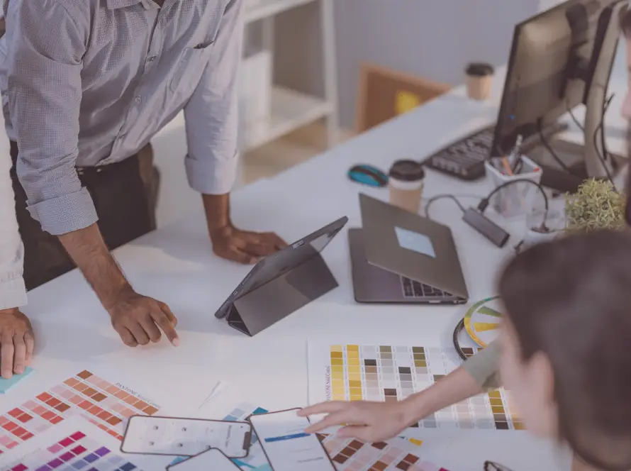 Four people gather around a white desk covered with colour swatches, charts, a mobile phone, a tablet, and a laptop. One person points at the swatches while others look on, suggesting a collaborative design or planning meeting in a modern office space.
