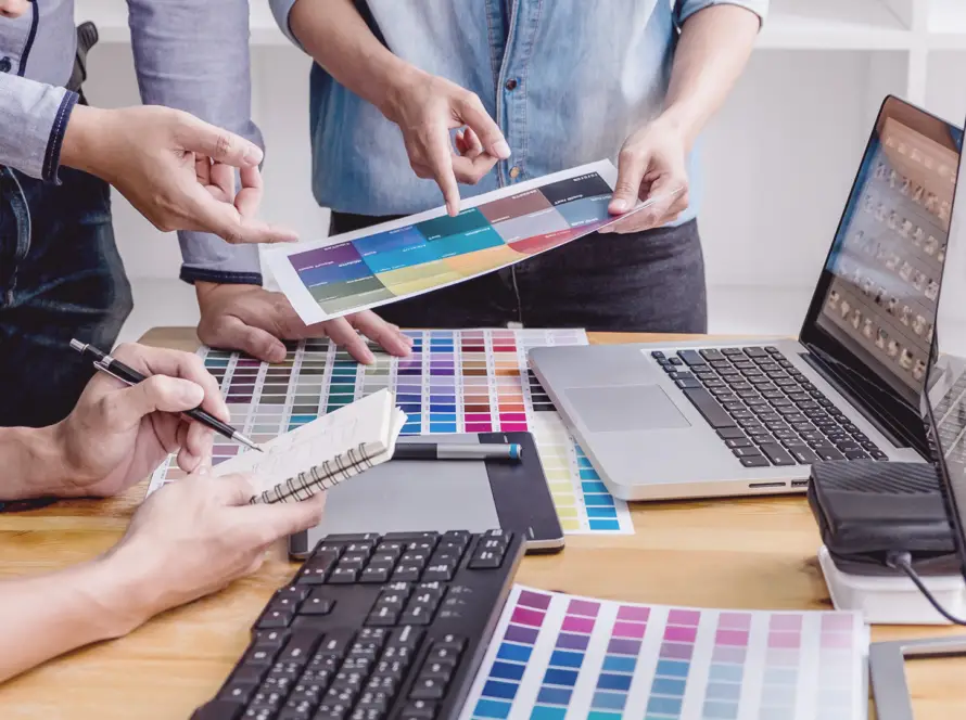 Three people collaborate at a desk covered with colour swatches, laptops, and a keyboard. One person writes in a notepad, another points at a colour chart, and the third holds a paper, suggesting a design or creative planning discussion.