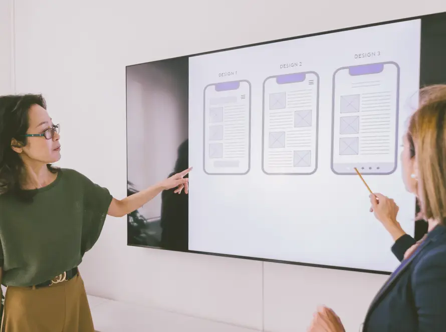 Two women in business attire stand in front of a large screen displaying three smartphone app wireframes labelled “DESIGN 1,” “DESIGN 2,” and “DESIGN 3.” Each woman points at a different design, appearing to discuss the options in an office setting.