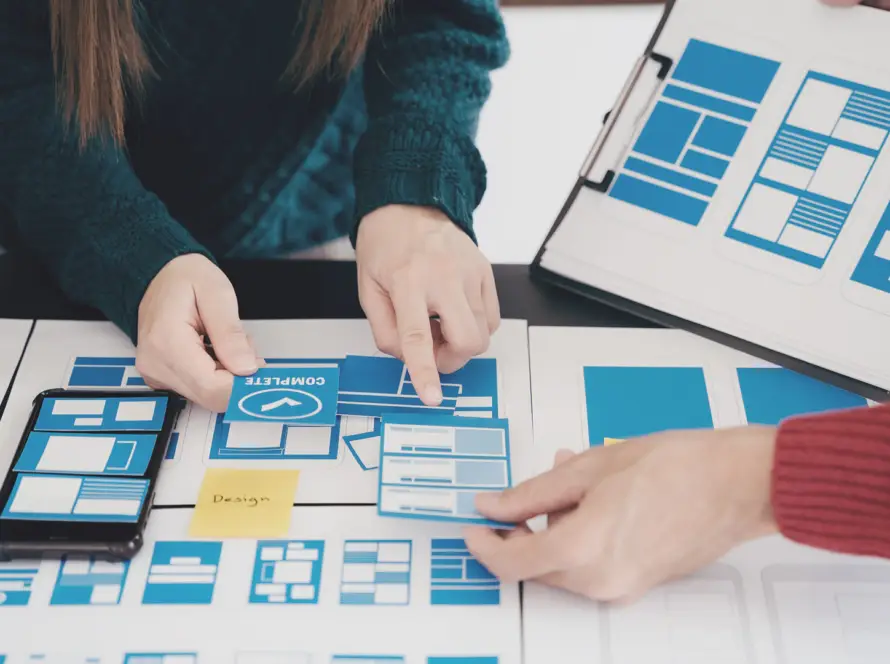 Three people collaborate at a table covered with blue and white website or app wireframes and mock-ups, pointing to layouts and discussing design. One person holds a clipboard with more layout options, emphasising teamwork in user interface design.
