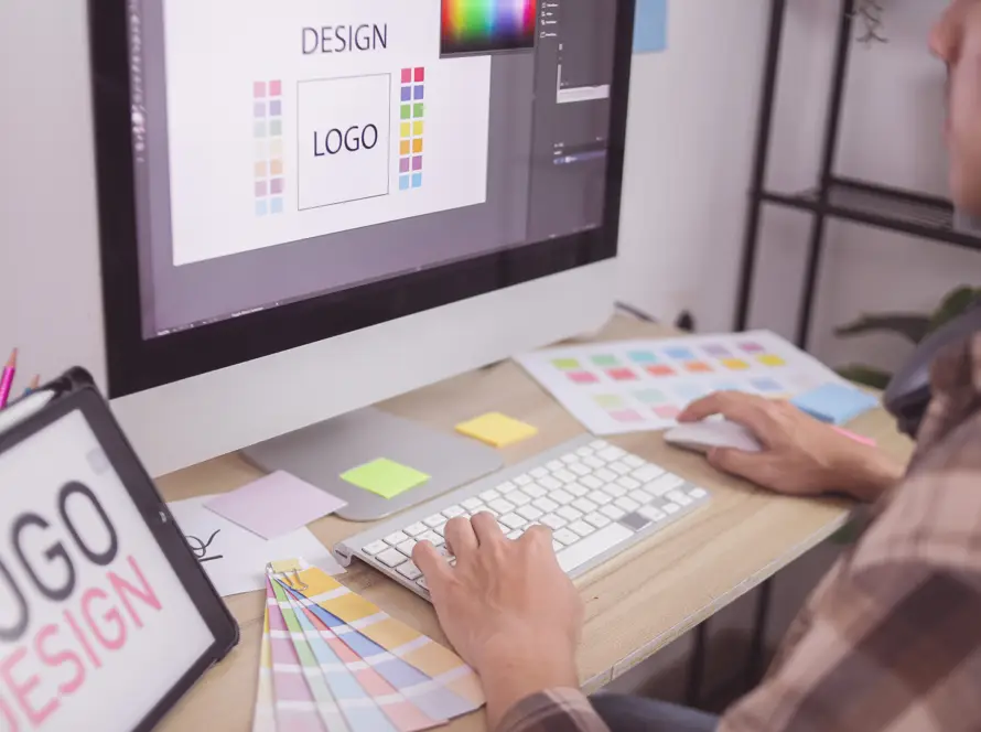 A person uses a desktop computer to design a logo, shown on the screen with various colour palettes. A colour swatch fan, sticky notes, and a tablet displaying LOGO DESIGN are on the desk. The person holds a keyboard and mouse.