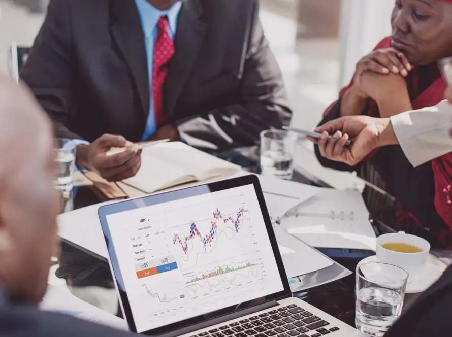 Four people in business attire sit around a glass table in discussion. A laptop displaying a colourful line graph with charts is in the foreground. Notebooks, papers, glasses of water, and a cup of tea are also on the table, indicating a work meeting.