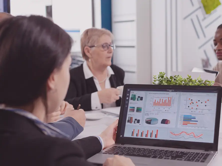 A group of four people sit at a table in an office, discussing. In the foreground, a laptop screen displays various colourful financial charts and graphs. One person takes notes, whilst others listen and talk, with papers and plants on the table.