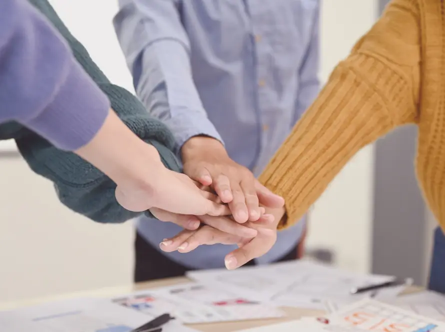 Four people stand around a table, stacking their hands together in a gesture of teamwork. They wear casual clothes: jumpers in green and yellow, and a blue button-up shirt. Papers and pens are scattered on the table beneath their hands.