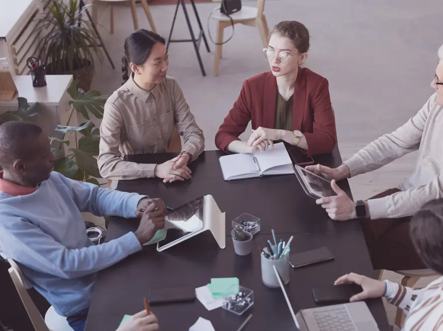 Six people of diverse backgrounds sit around a table in an office. They discuss, take notes, and use digital devices. The group includes men and women, plants decorate the space, and notebooks, tablets, and papers are spread on the table.