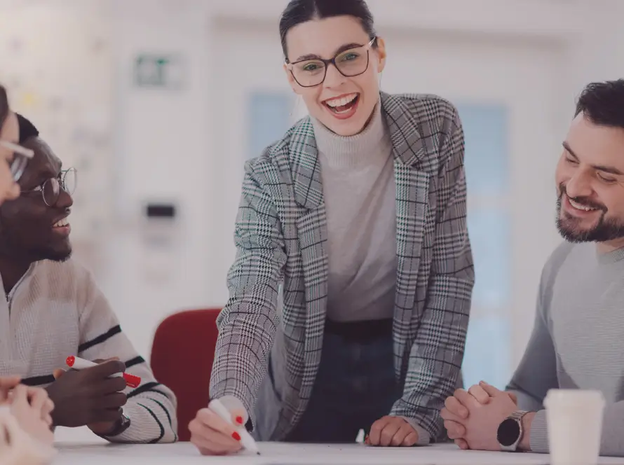A woman in a checked blazer and glasses stands smiling at a table with three colleagues. She is holding a marker and appears to be leading a discussion or presentation. The group looks engaged and happy in a bright, modern office setting.