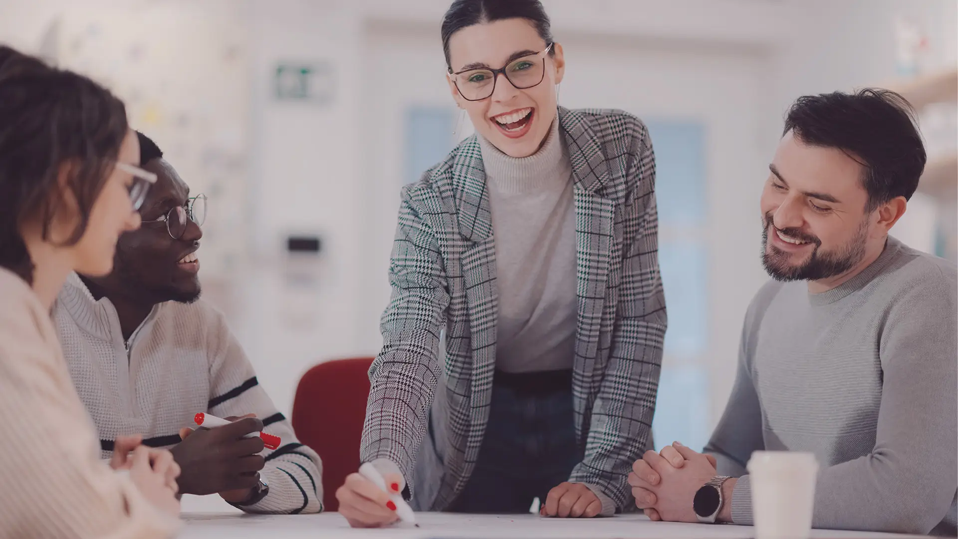 A woman in a checked blazer and glasses stands smiling at a table with three colleagues. She is holding a marker and appears to be leading a discussion or presentation. The group looks engaged and happy in a bright, modern office setting.