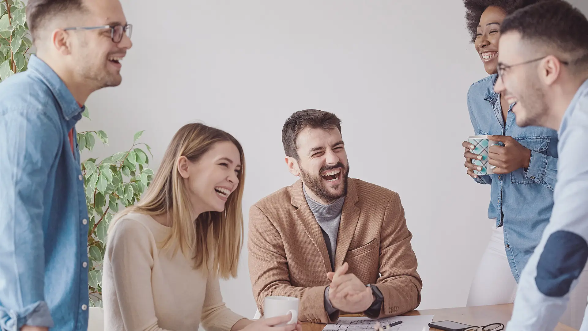 Five coworkers, three men and two women, stand around a desk laughing together. They appear relaxed and friendly, holding mugs and wearing casual business attire. A potted plant is in the background against a plain, bright wall.