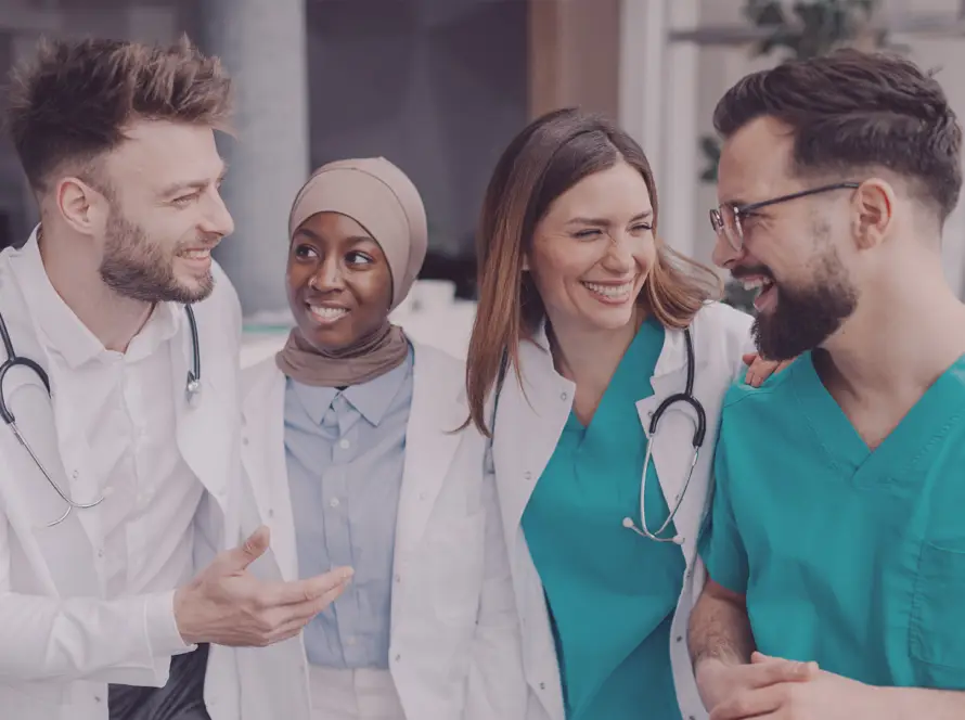 Four diverse medical professionals, two women and two men, stand together smiling and talking. They wear medical attire—white coats and teal scrubs—with stethoscopes around their necks, showing camaraderie and teamwork in a bright, modern setting.