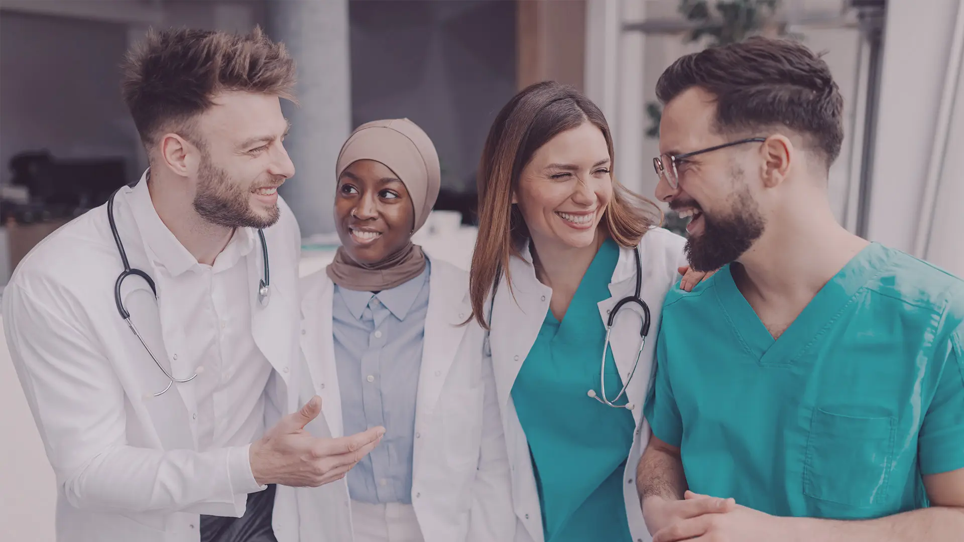 Four diverse medical professionals, two women and two men, stand together smiling and talking. They wear medical attire—white coats and teal scrubs—with stethoscopes around their necks, showing camaraderie and teamwork in a bright, modern setting.