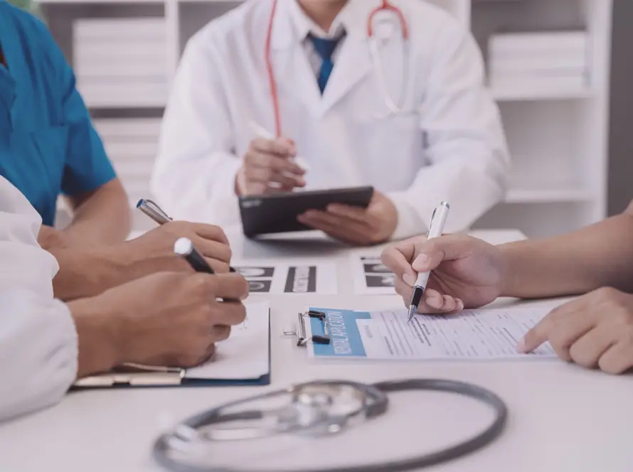 Four healthcare professionals in scrubs and lab coats sit at a table, writing on clipboards and discussing documents. A stethoscope lies on the table, and one person holds a tablet. The atmosphere is collaborative and focused.