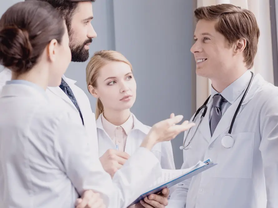 Four doctors in white coats stand together indoors, having a discussion. One doctor holds a clipboard and gestures as they talk. Another doctor, wearing a stethoscope, smiles. The group appears engaged in a professional conversation near a window.