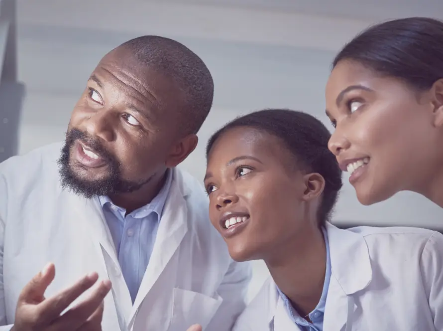 Three people wearing white lab coats closely examine something off-screen. They appear engaged and are smiling. The background is blurred, focusing attention on their attentive expressions and collaborative interaction, suggesting a scientific or medical setting.
