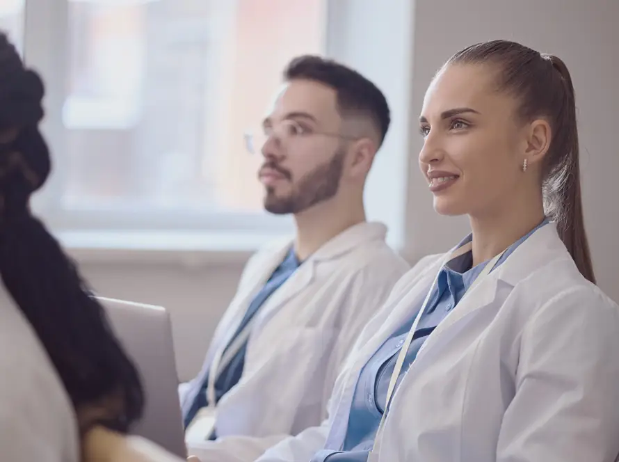Several young adults in white lab coats sit attentively in a bright classroom. The focus is on a woman with a ponytail smiling, and a man with glasses beside her. Both appear engaged, suggesting a medical or healthcare educational setting.